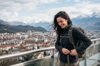 Jeune femme souriante ajustant son sac sur un balcon avec vue sur Grenoble et les Alpes